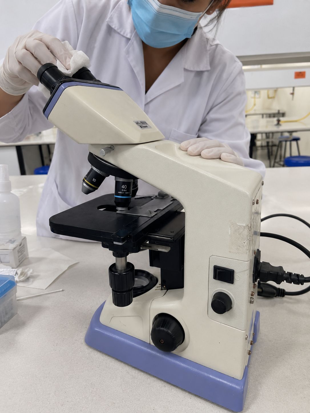 Technician cleaning a microscope eyepiece with a cloth, wearing gloves and a mask