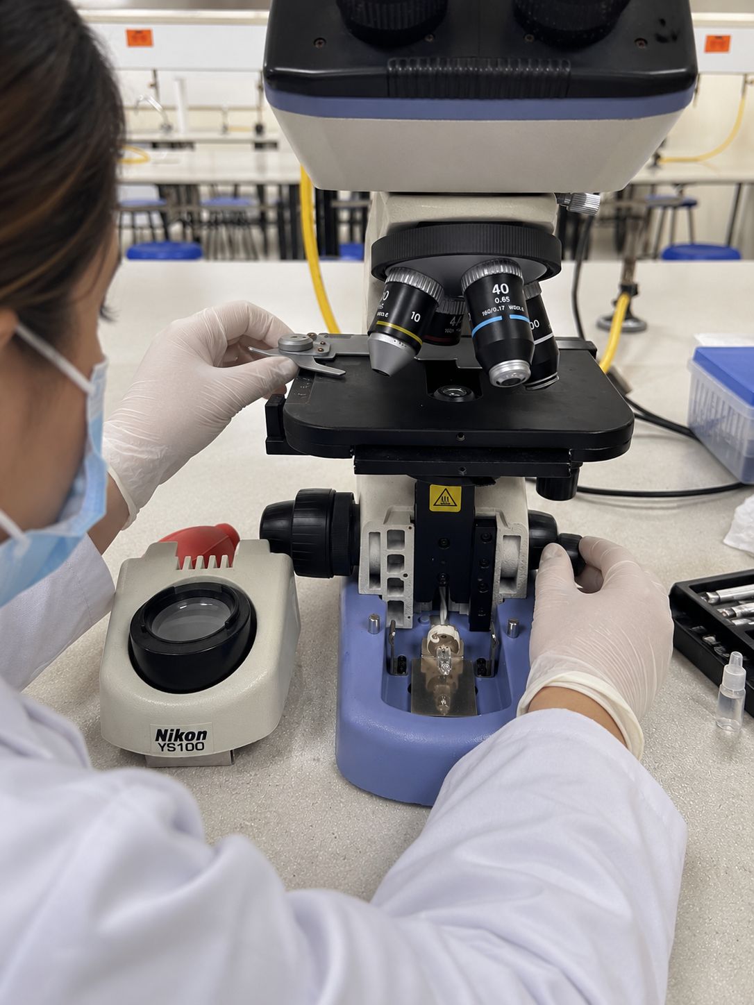 Technician wearing gloves and a mask adjusting the stage and focus of a Nikon microscope